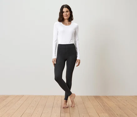 Une femme se tient sur un plancher en bois, portant un t-shirt blanc à manches longues et des leggings foncés.
