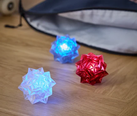 Trois boules lumineuses reposent sur un plancher en bois : une blanche, une bleue et une rouge. Derrière elles se trouvent un sac à dos et une planche à roulettes.