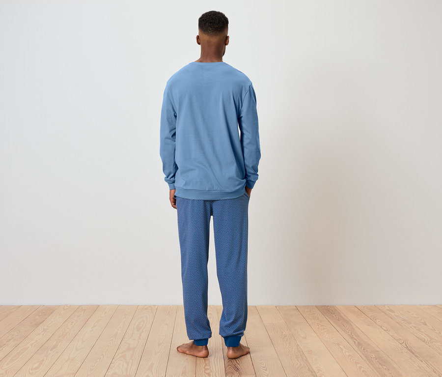 Un homme vu de dos portant un pyjama bleu se tient sur un plancher en bois devant un mur blanc.