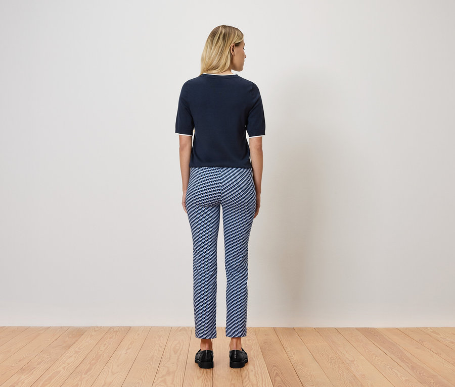 Une femme blonde se tient dos à la caméra, portant un haut à manches courtes bleu marine et un pantalon rayé bleu et blanc. Elle se tient sur un plancher en bois.
