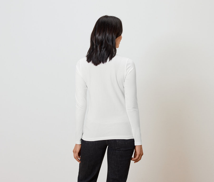 Une femme aux cheveux courts et foncés se tient dos à la caméra, portant un haut blanc à manches longues et un jean noir.