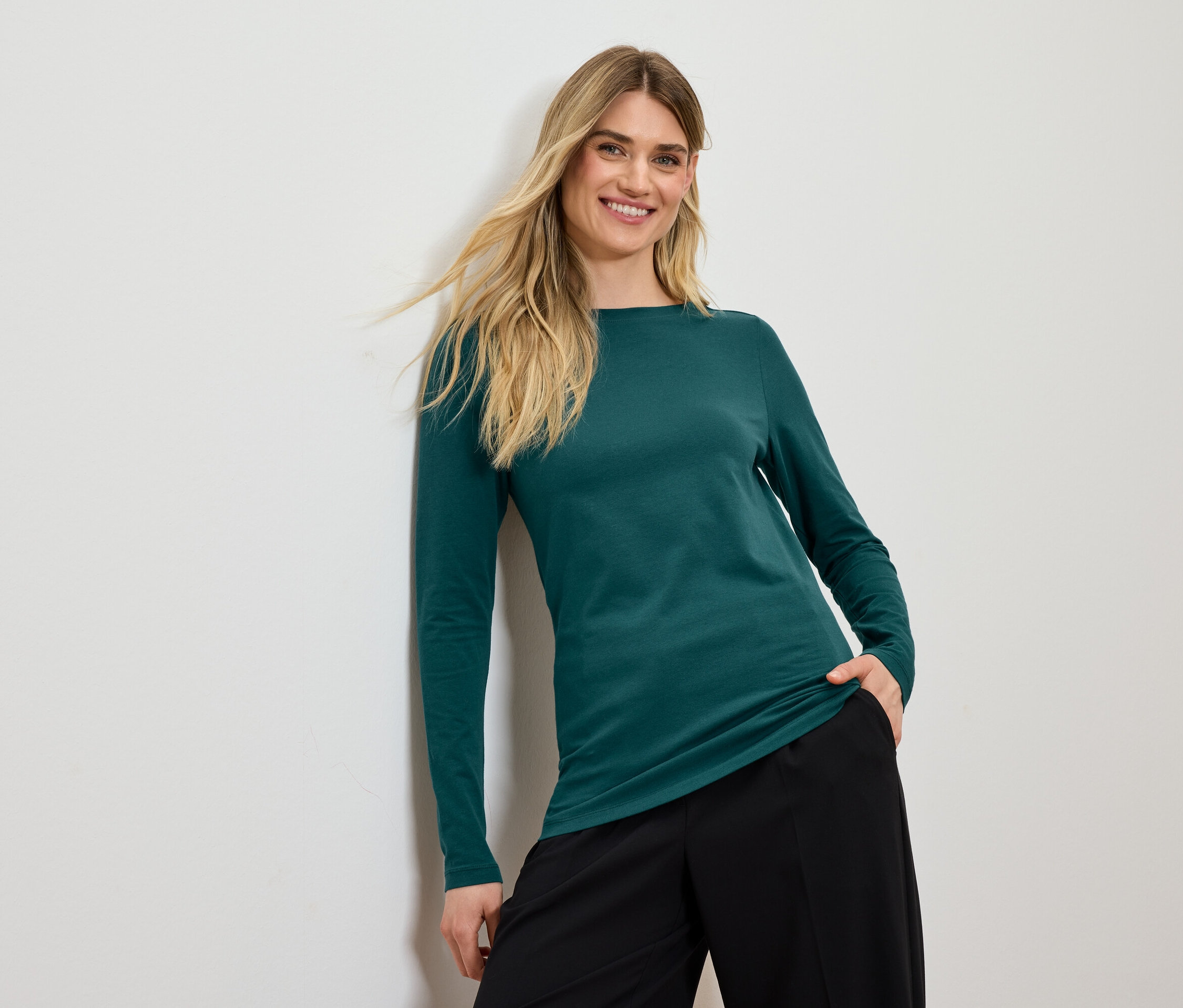 Une femme souriante aux cheveux blonds pose dans un haut vert à manches longues et un pantalon noir.