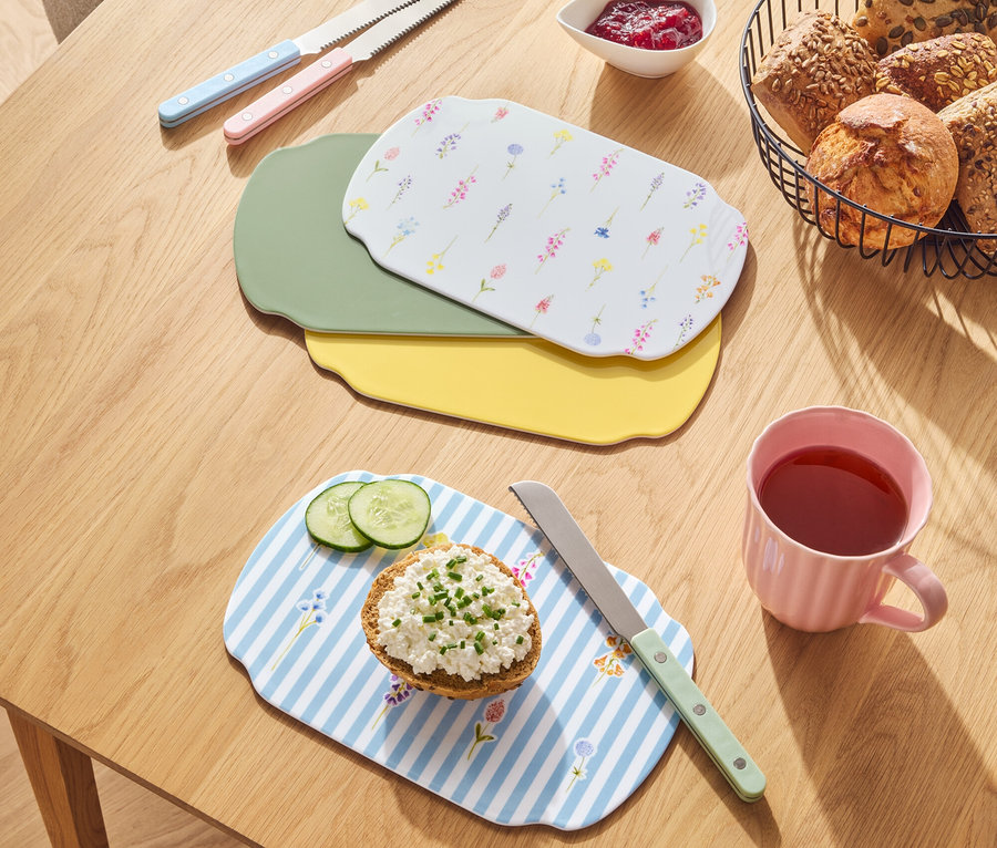 Sur une table en bois se trouvent des pains dans un panier, de la confiture, des couteaux et des planches à découper de différentes couleurs.