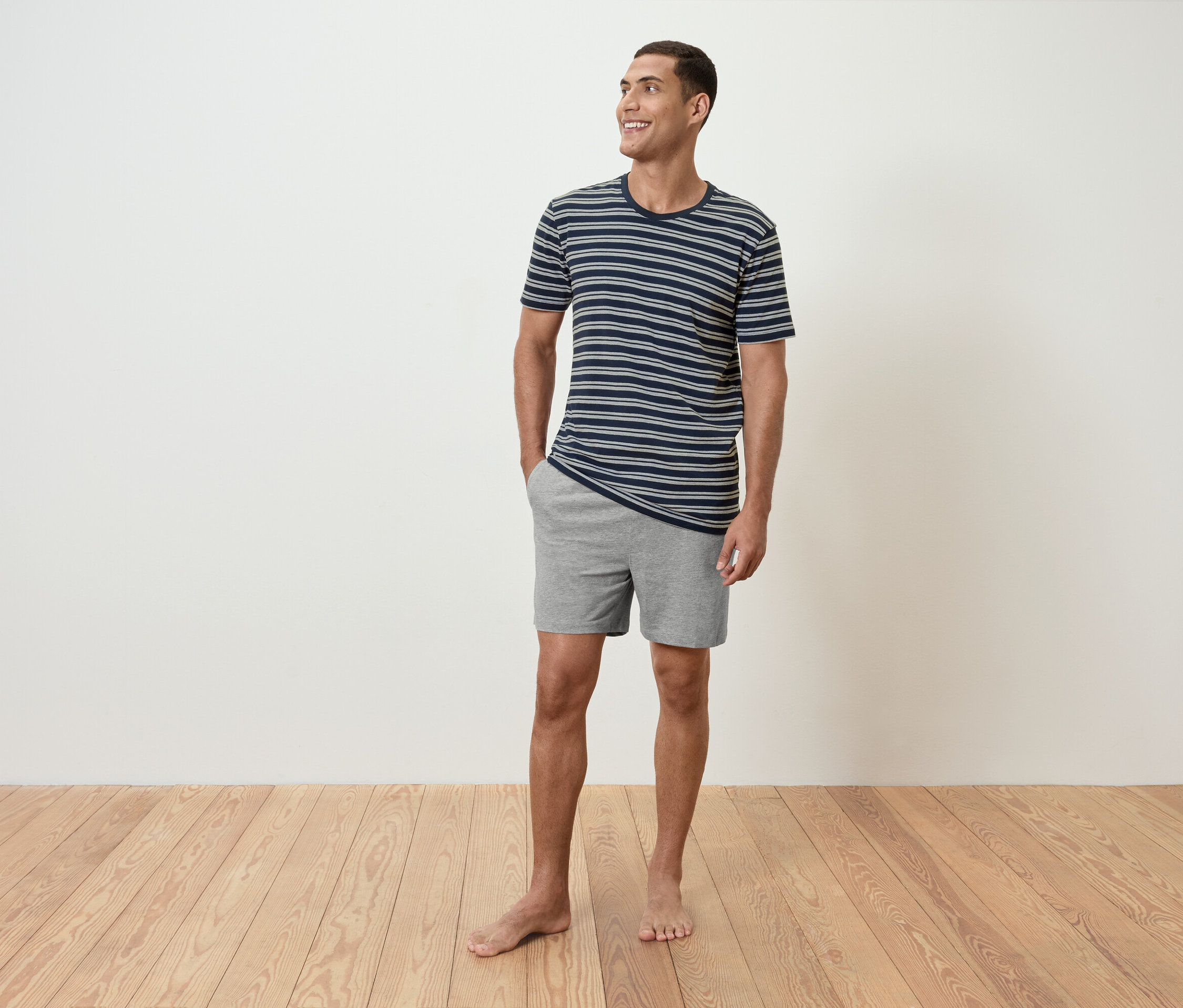 Un homme se tient pieds nus sur un plancher en bois, portant un t-shirt rayé et un short gris.