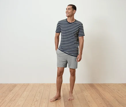 Un homme se tient pieds nus sur un plancher en bois, portant un t-shirt rayé et un short gris.