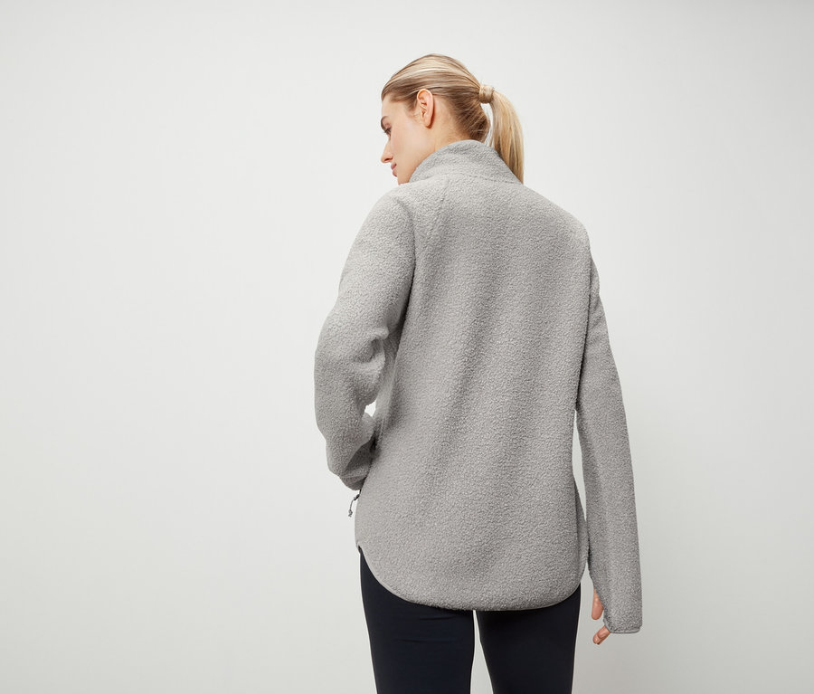 De dos, une femme aux cheveux blonds attachés en queue de cheval porte un gilet en polaire gris et un pantalon bleu marine.
