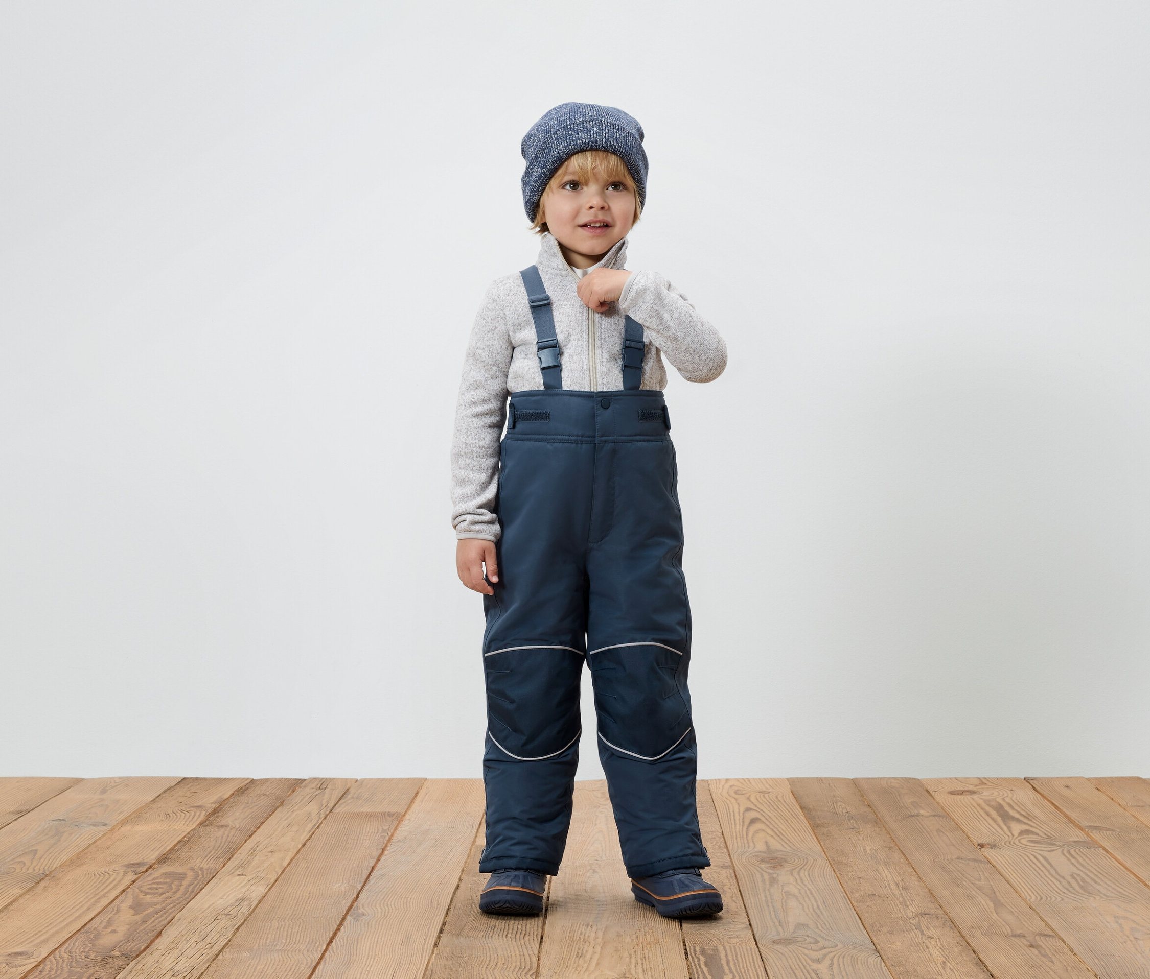 Un garçon en combinaison d'hiver bleue avec des bretelles et un bonnet en tricot se tient sur un plancher en bois et ferme sa veste.