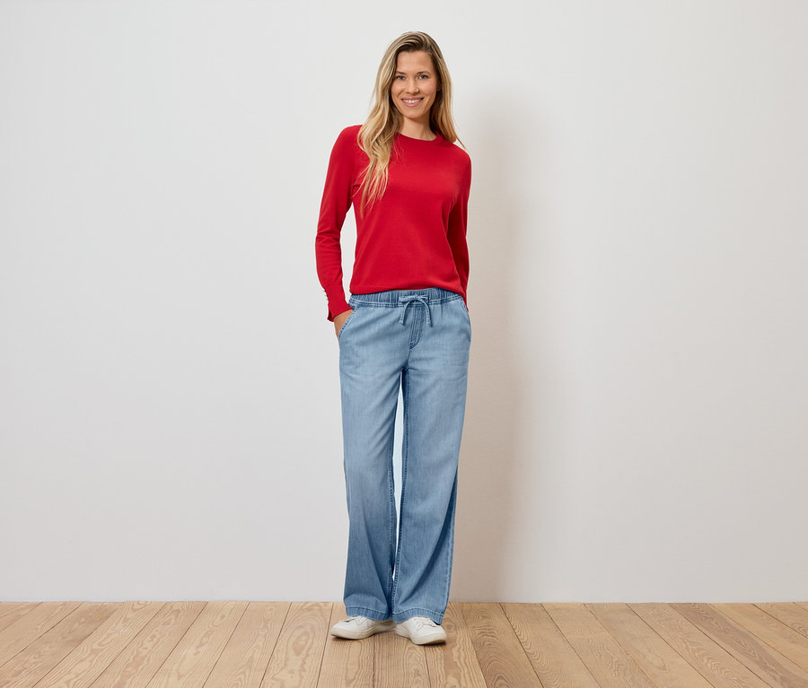 Une femme portant un pull rouge et un pantalon évasé bleu clair se tient sur un plancher en bois.