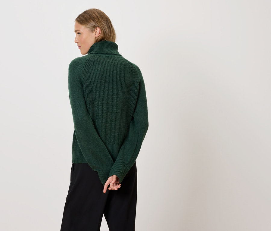 Une femme blonde, vue de dos, porte un pull à col roulé vert et un pantalon noir.
