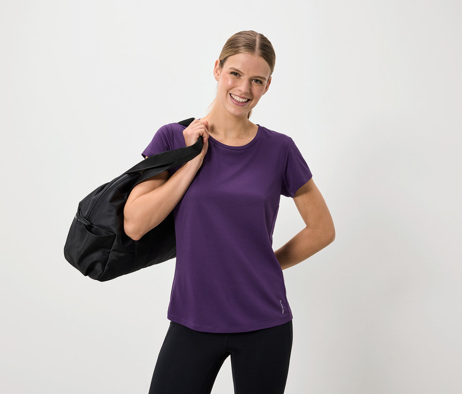 Femme souriante portant un t-shirt violet et un legging noir avec un sac noir sur l'épaule.