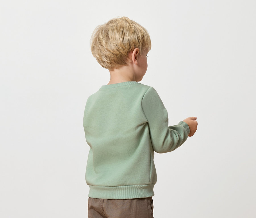 Un garçon blond, vêtu d'un pull vert et d'un pantalon marron, se tient dos à l'appareil photo.