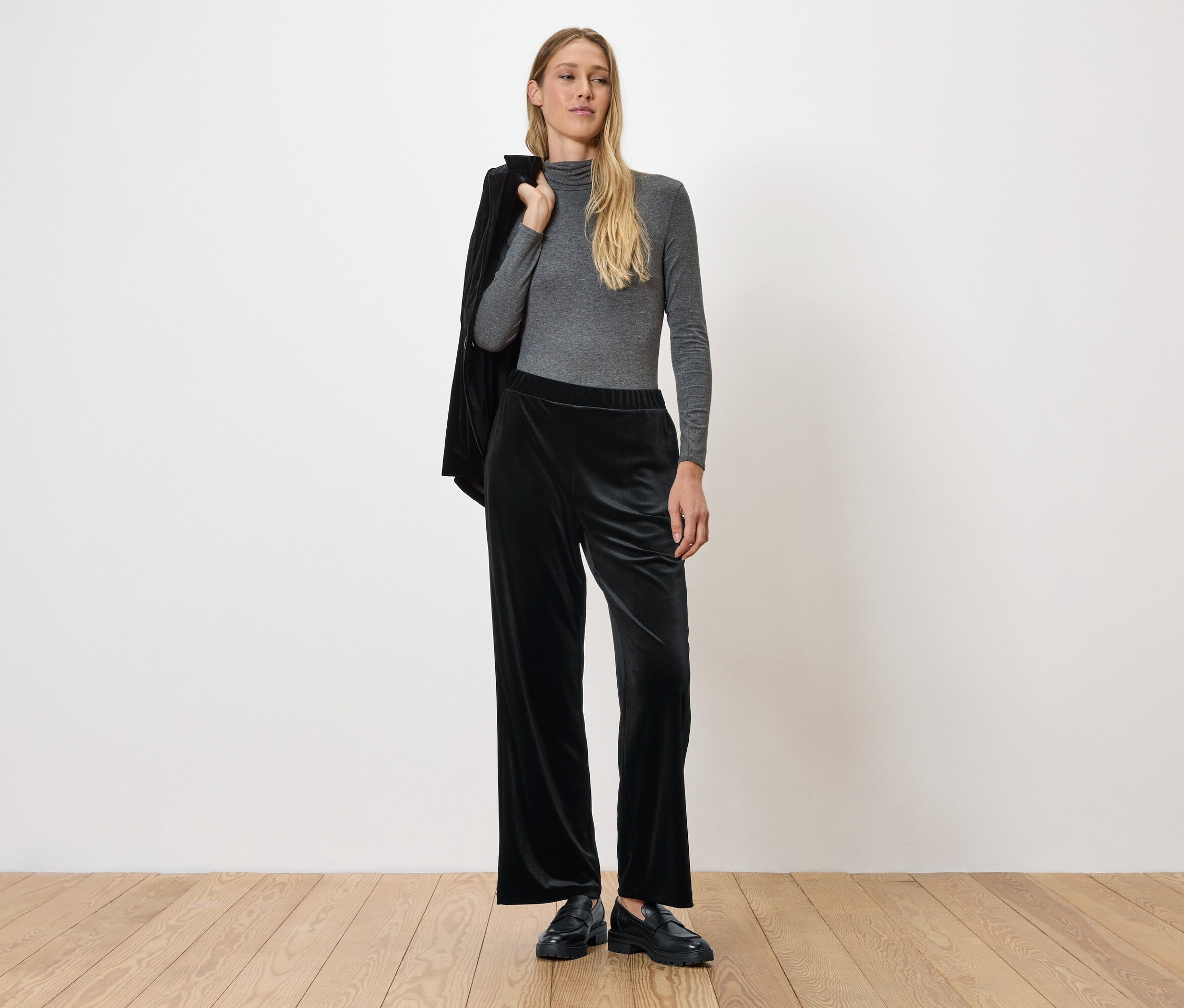 Une femme pose en haut gris à manches longues et pantalon en velours noir, une veste sur l'épaule.