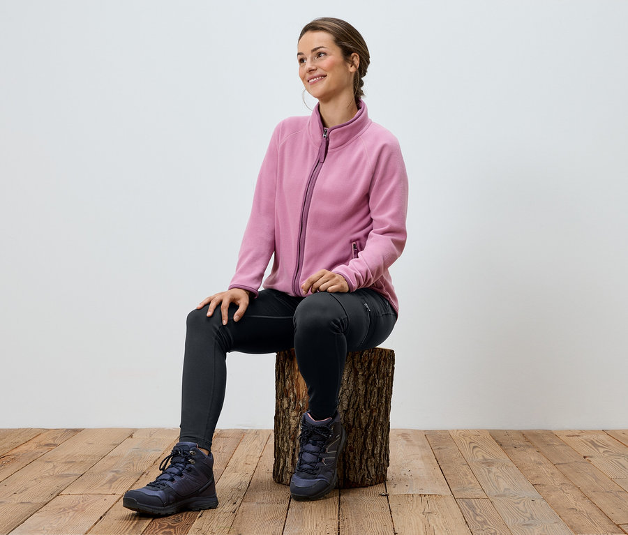Une femme est assise sur un tabouret en bois, portant une veste en polaire rose, un legging noir et des chaussures de randonnée.