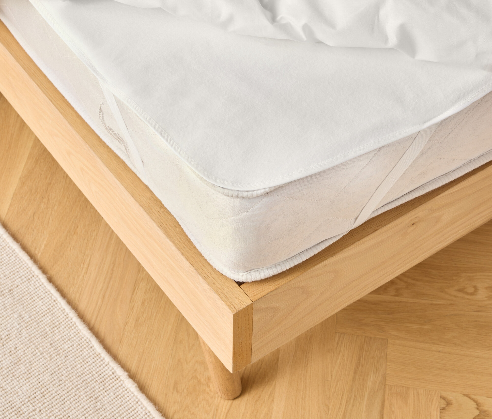 Vue d'un cadre de lit en bois clair avec un matelas blanc et une couette blanche. Un tapis beige est partiellement visible.