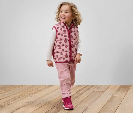 Une fillette souriante aux cheveux bouclés, vêtue d'un gilet rose à pois, d'un pantalon rose et de chaussures roses, se tient debout sur un plancher en bois.