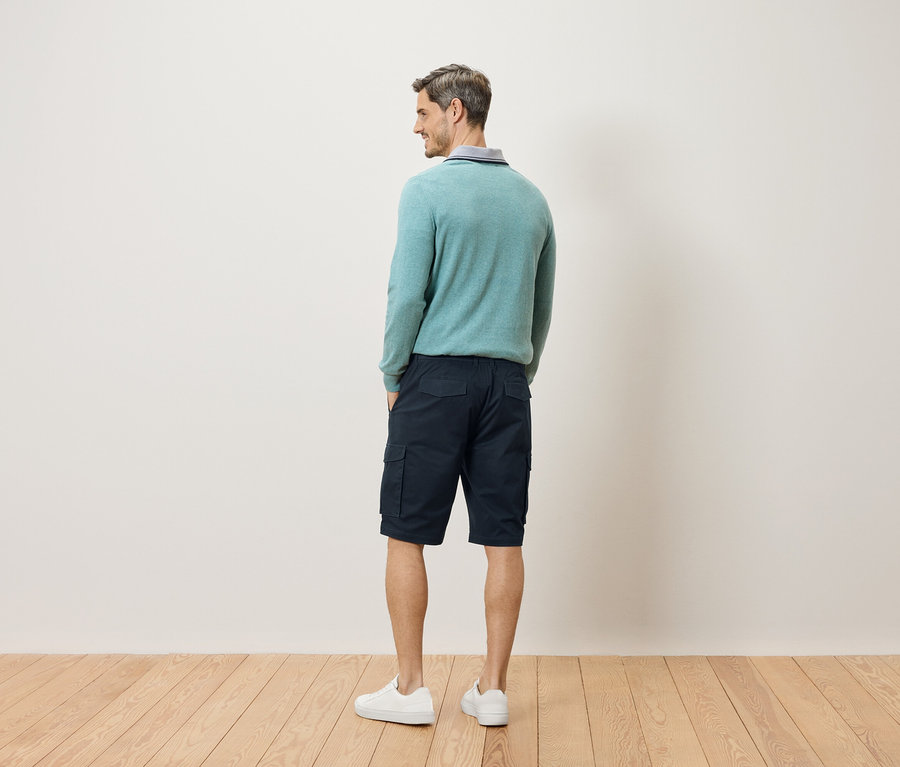 Un homme se tient dos à la caméra, portant un pull bleu clair, un short cargo et des baskets blanches.
