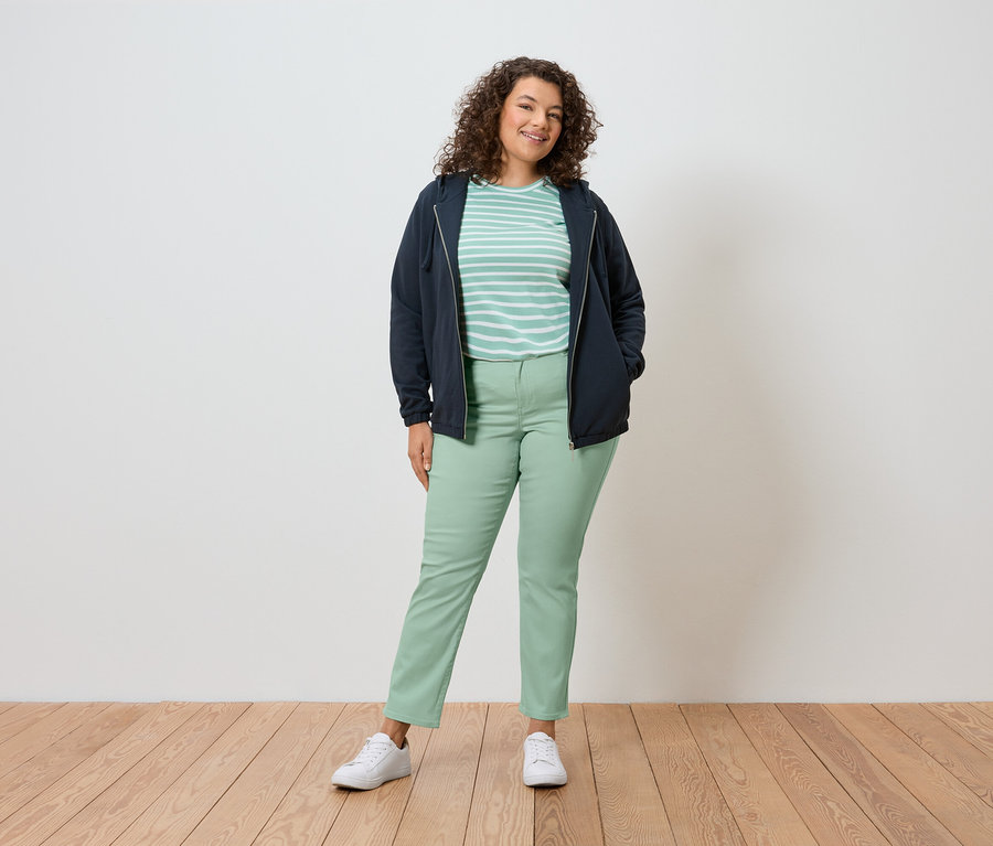 Une femme aux cheveux bouclés se tient devant un mur, portant une veste bleu marine, un t-shirt rayé, un pantalon vert et des chaussures blanches.
