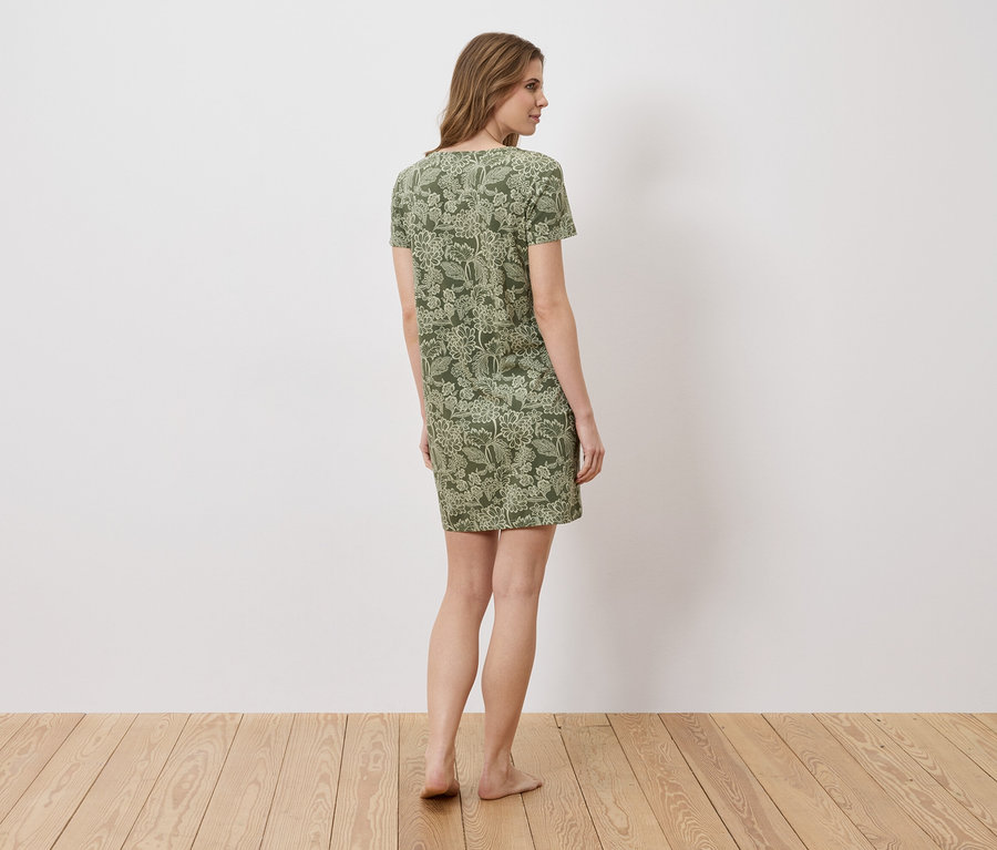 Une femme se tient pieds nus dans une robe verte à manches courtes et à motifs floraux devant un mur blanc.