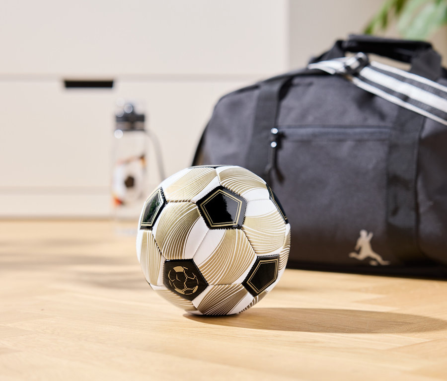 Un ballon de foot doré est posé sur un plancher en bois, avec un sac de sport et une bouteille d'eau en arrière-plan.