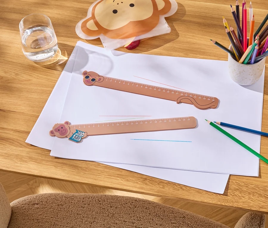 Deux règles marron avec des motifs de singe sont posées sur une feuille de papier, à côté d'un verre d'eau, de crayons et d'une trousse en forme de singe.