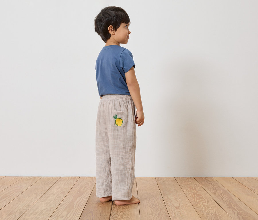 Un garçon se tient pieds nus sur un plancher en bois, portant un t-shirt bleu et un long pantalon marron clair avec un citron sur la poche.