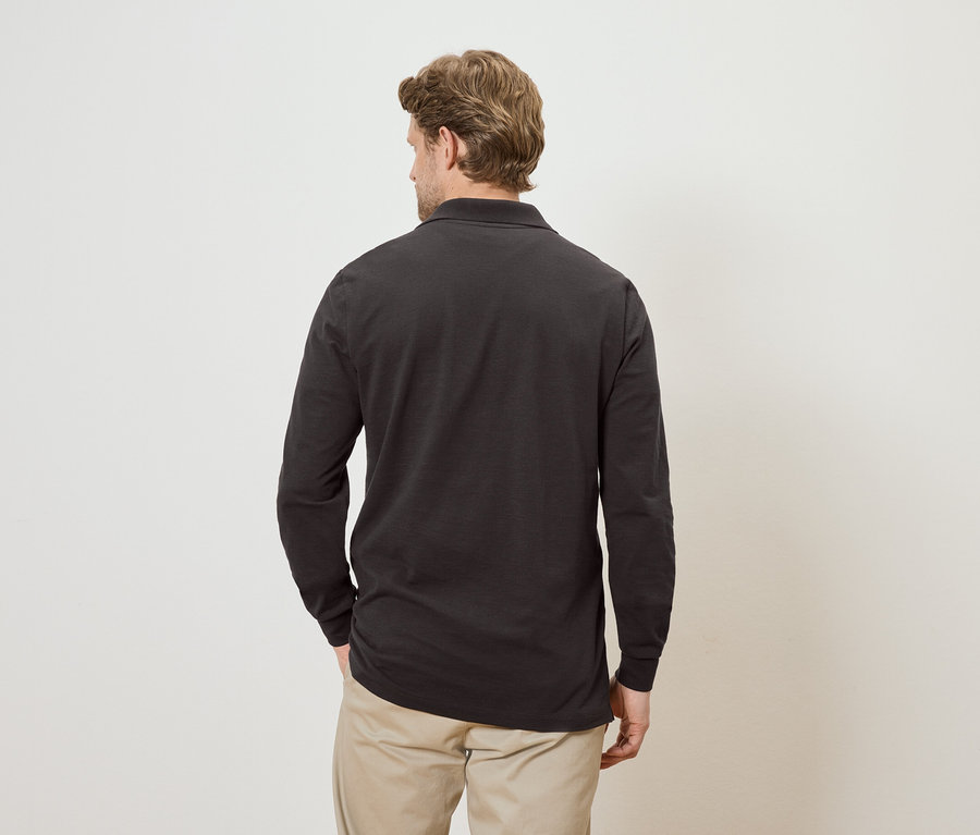 Vue de dos d'un homme blond portant un polo à manches longues gris foncé et un pantalon beige sur un fond crème.