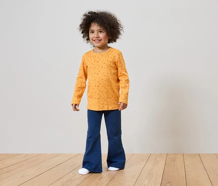 Une fillette souriante aux cheveux bouclés porte un t-shirt orange à manches longues à motifs et un pantalon bleu patte d'eph.