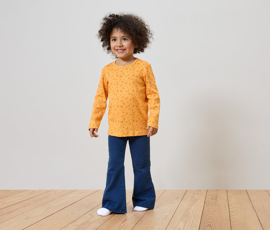 Une fillette souriante aux cheveux bouclés porte un t-shirt orange à manches longues à motifs et un pantalon bleu patte d'eph.