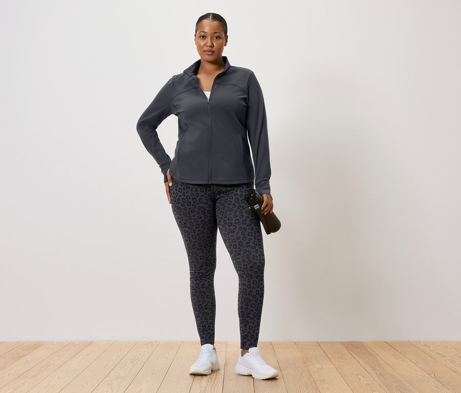 Femme en veste de survêtement foncée et leggings à motif léopard tenant une bouteille d'eau.