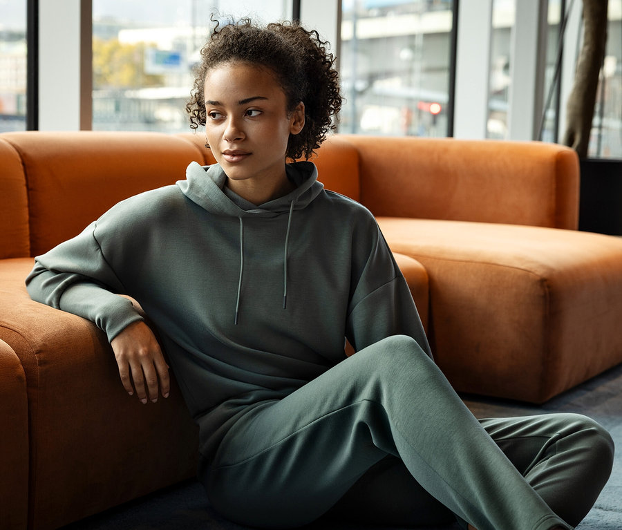 Une femme est assise par terre, appuyée contre un canapé orange, et porte un sweat à capuche gris et un pantalon de jogging.