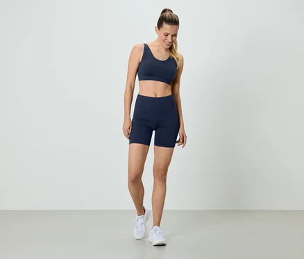 Femme en soutien-gorge de sport bleu marine et short de cycliste, avec des baskets blanches, debout devant un fond blanc.