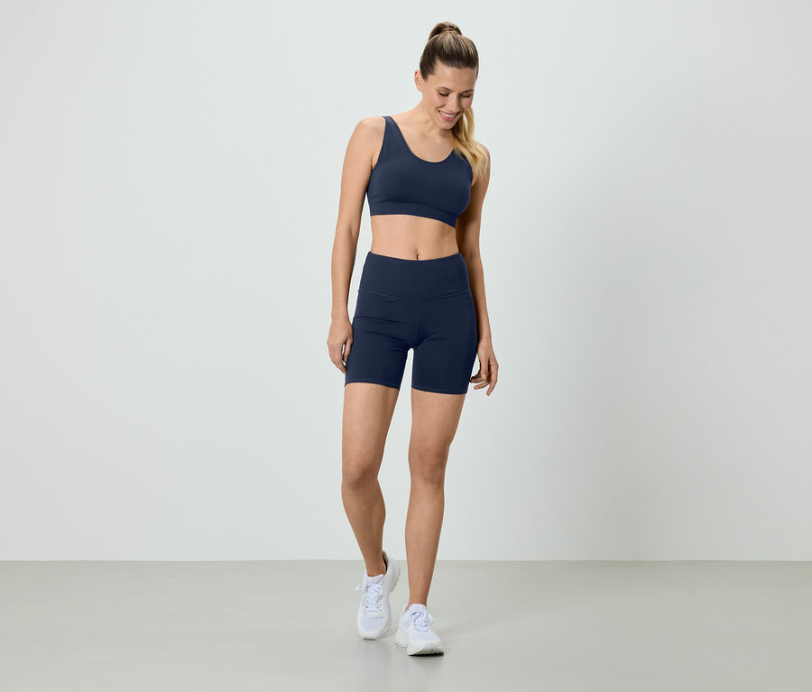Femme en soutien-gorge de sport bleu marine et short de cycliste, avec des baskets blanches, debout devant un fond blanc.