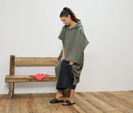 Femme changeant un pantalon noir sous un poncho à capuche vert sur un banc en bois, à côté d'elle des vêtements rouges.