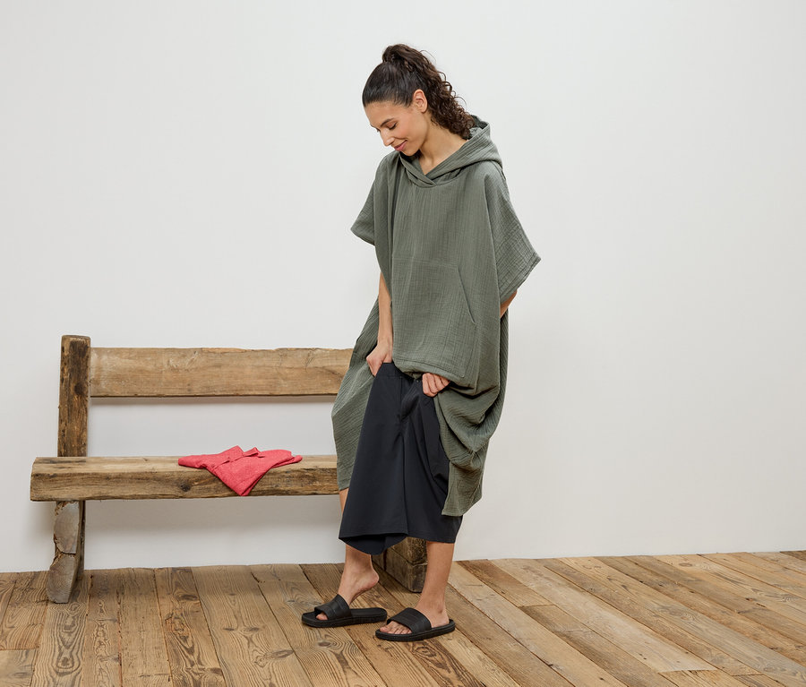 Femme changeant un pantalon noir sous un poncho à capuche vert sur un banc en bois, à côté d'elle des vêtements rouges.