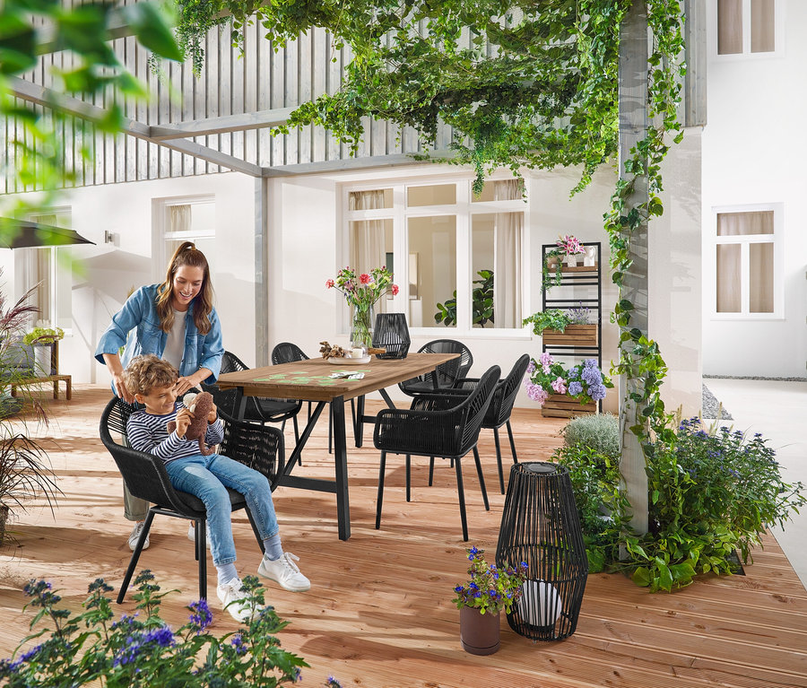 Une mère et son enfant sont assis à une table de jardin extensible avec un cadre rustique d'environ 200 à 250 cm. Une petite et une grande Lanterne solaire avec ruban en tissu sont posées sur le sol. À l'arrière-plan, Mur végétal avec étagères flexiblement accrochables « Elin ».