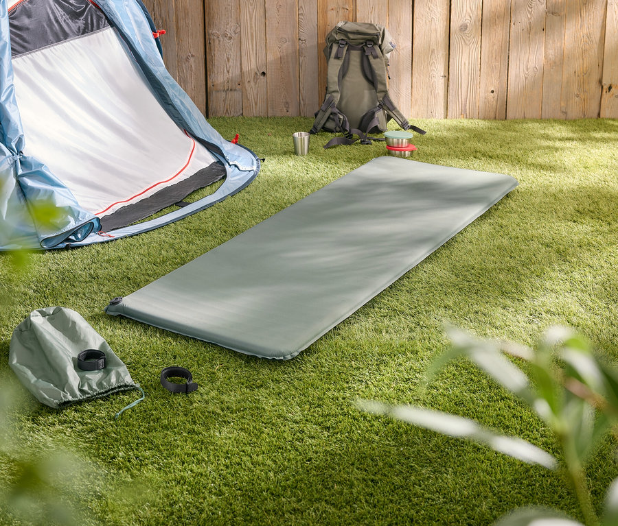Une tente bleue, un tapis de sol gris, une tasse argentée et un sac à dos gris sont posés sur une pelouse verte devant une clôture en bois.