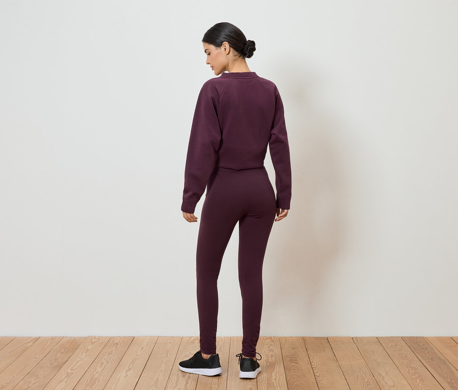 Vue arrière d'une femme portant un chignon, un survêtement bordeaux et des baskets noires, se tenant sur un plancher en bois devant un mur blanc.
