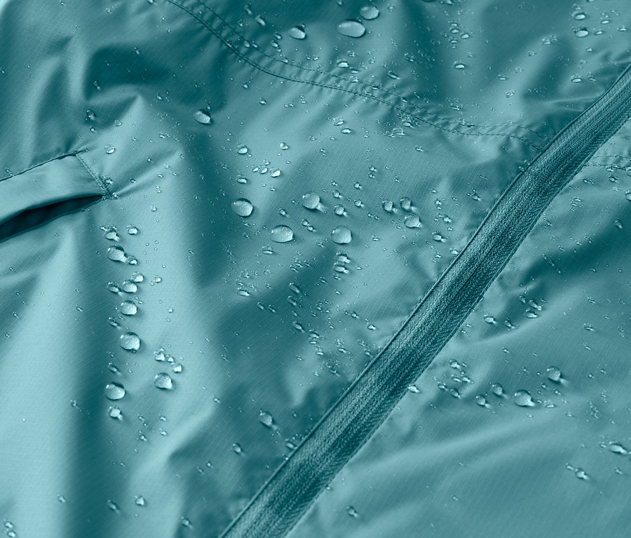 Gros plan d'une veste turquoise imperméable avec une fermeture éclair et une poche, sur laquelle reposent des gouttes d'eau.