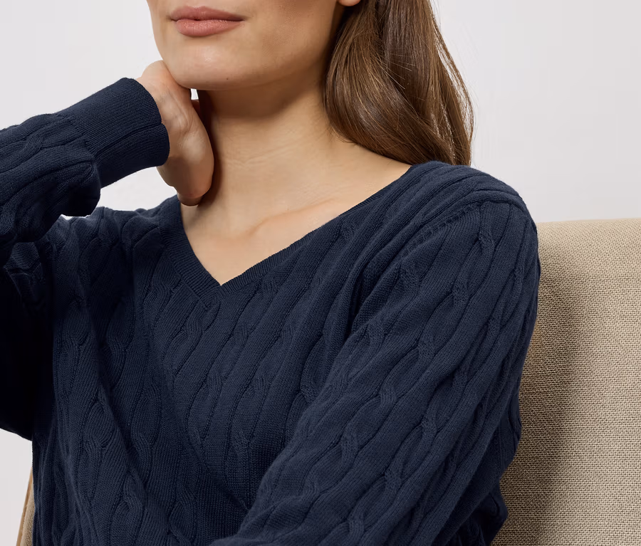 Une femme aux cheveux bruns porte un pull bleu marine à col en V et touche son cou.
