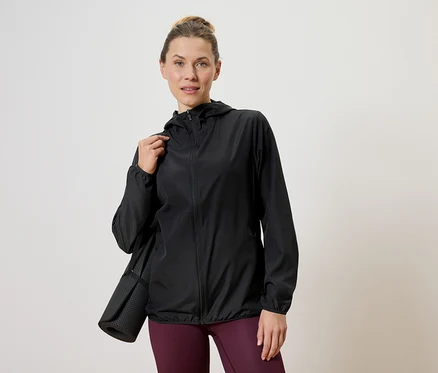 Femme portant une veste noire avec un tapis de yoga sur l'épaule, sur un fond neutre.