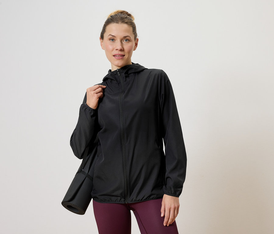 Femme portant une veste noire avec un tapis de yoga sur l'épaule, sur un fond neutre.
