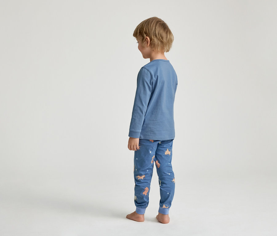 Un jeune enfant est debout, de dos, vêtu d'un pyjama bleu. Le haut à manches longues est bleu uni et le bas de pyjama est bleu avec un motif de chiens de dessin animé. L'enfant est pieds nus.