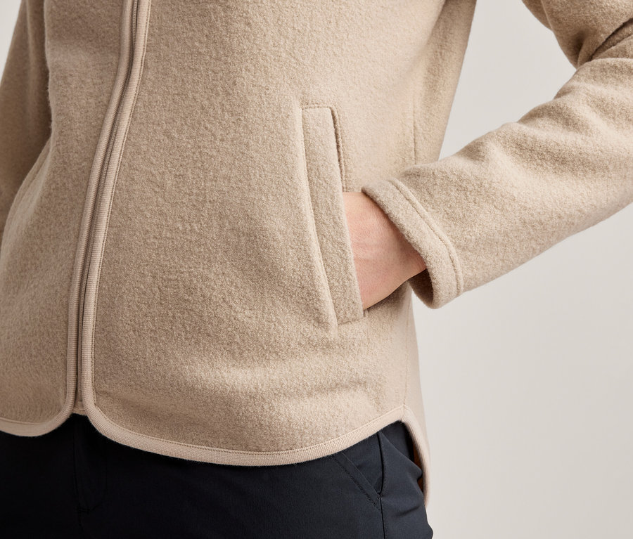 Gros plan d'une personne portant une veste polaire beige avec une fermeture éclair visible et une main dans une poche latérale. Un pantalon foncé est visible sous la veste.