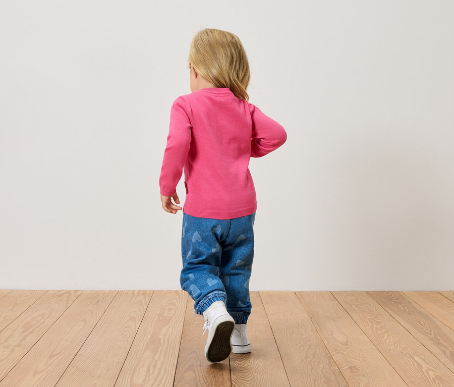 Petit enfant blond vu de dos, portant un pull rose, un jean bleu avec des cœurs et des chaussures blanches, marchant sur un plancher en bois.