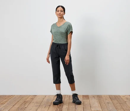 Une femme se tient devant un mur blanc, portant un t-shirt vert, un pantalon capri noir et des chaussures de randonnée noires.