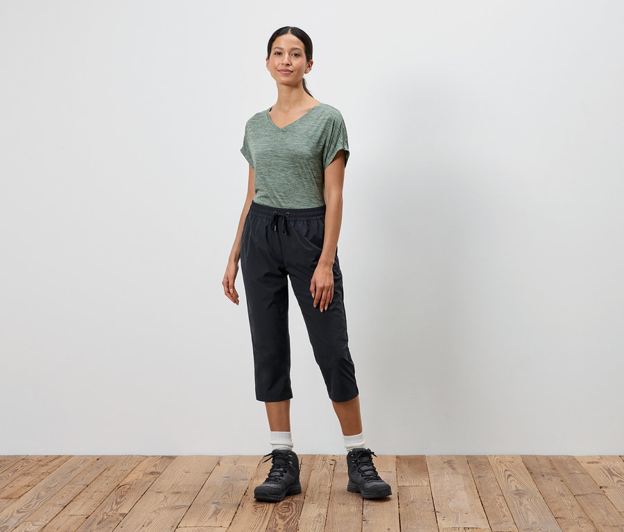 Une femme se tient devant un mur blanc, portant un t-shirt vert, un pantalon capri noir et des chaussures de randonnée noires.
