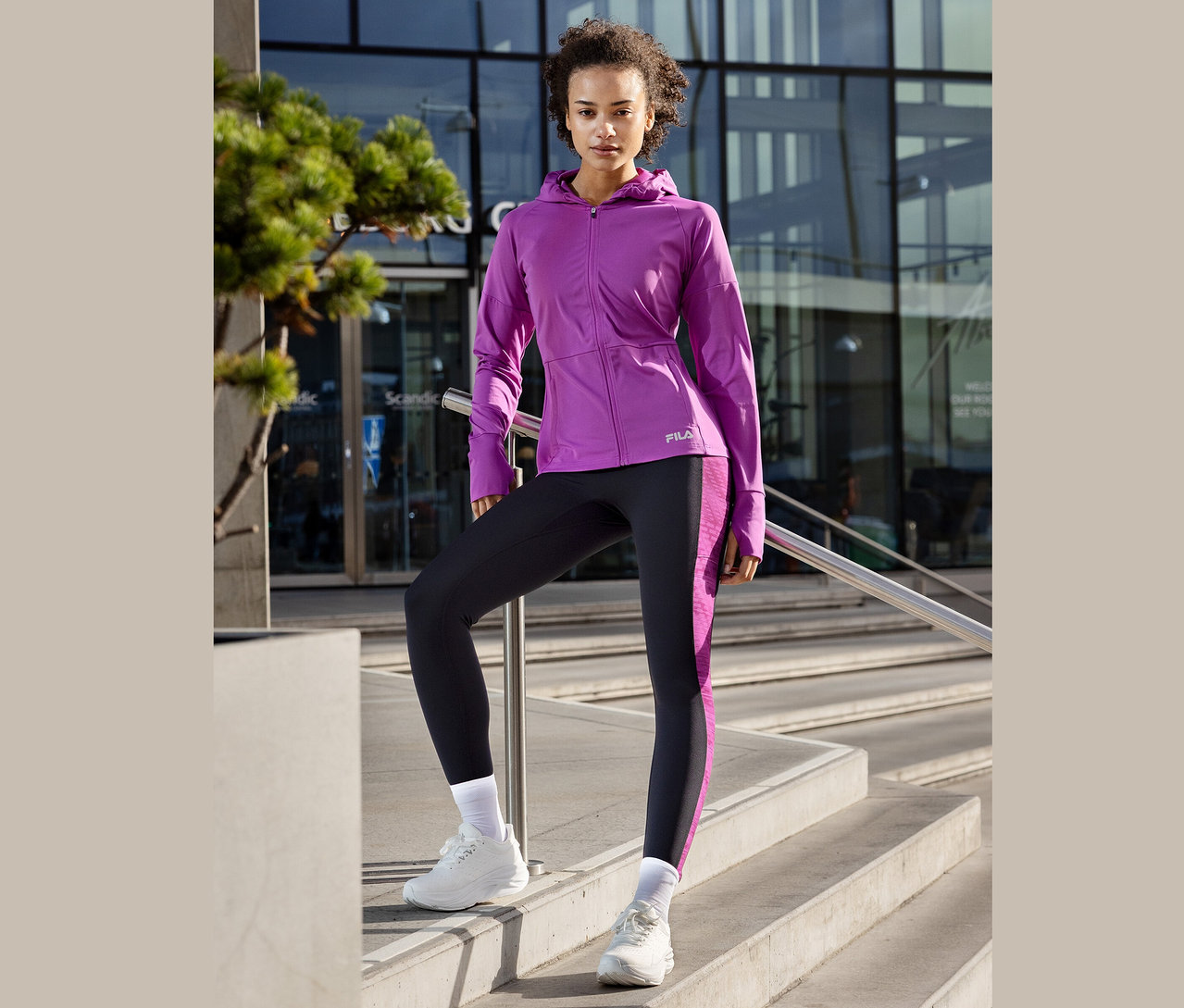 Femme posant sur un escalier, portant une veste Fila violette, un legging avec des rayures violettes et des baskets blanches.