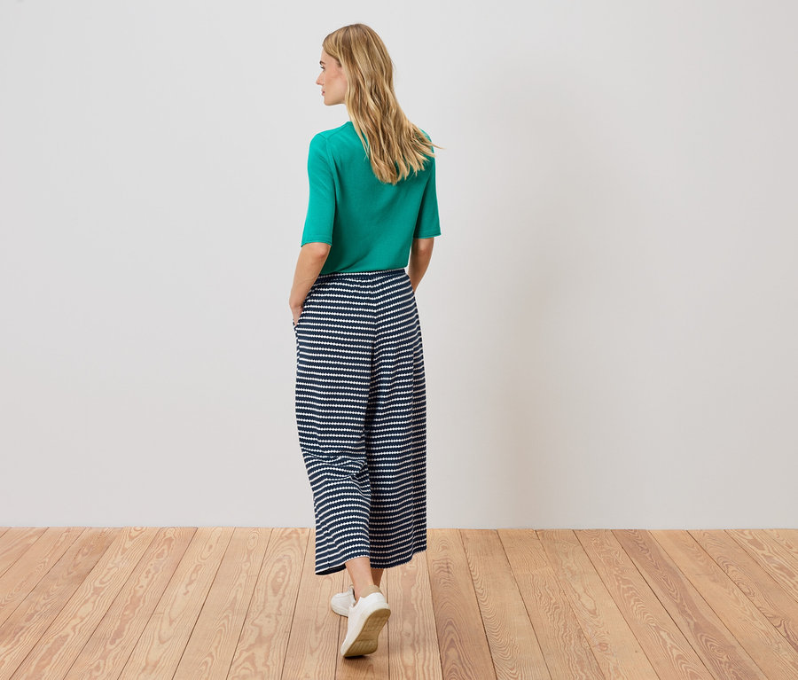 Femme de dos portant un haut vert et un pantalon rayé sur un plancher en bois.