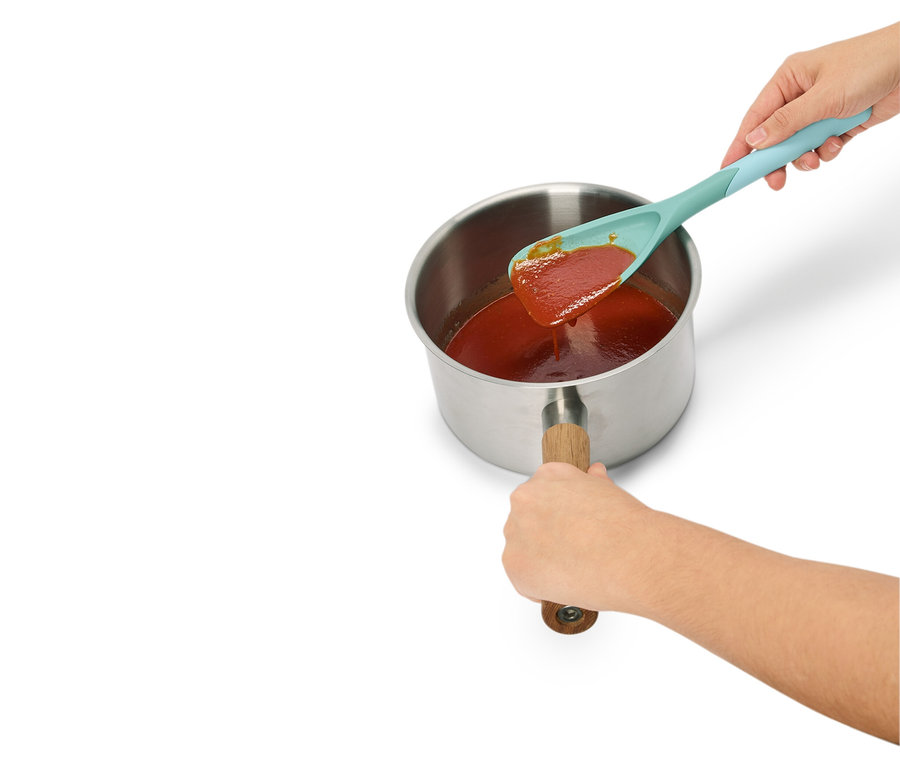 Une main tient une cuillère turquoise avec de la sauce rouge au-dessus d'une casserole en acier avec une poignée en bois.