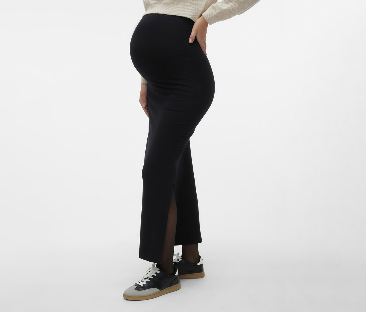 Femme enceinte portant une jupe maxi MAMALICIOUS «
MLEMMA TUBE
» avec une fente latérale, des collants foncés et des baskets sur un fond neutre.